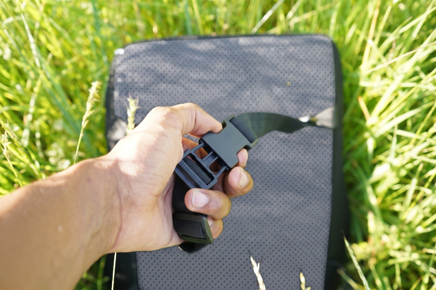 Eine Hand hält den Gurtverschluss des Kissens, das Kissen liegt dahinter im Gras.